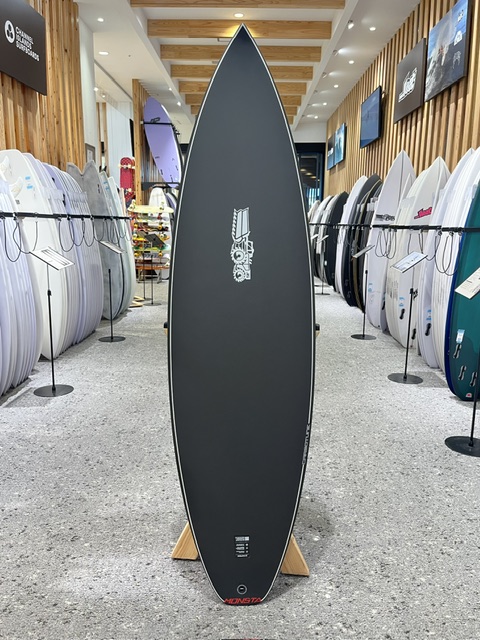 Long dark gray surfboard on a wooden stand in a bright surf shop showroom, flanked by racks of boards along both walls.