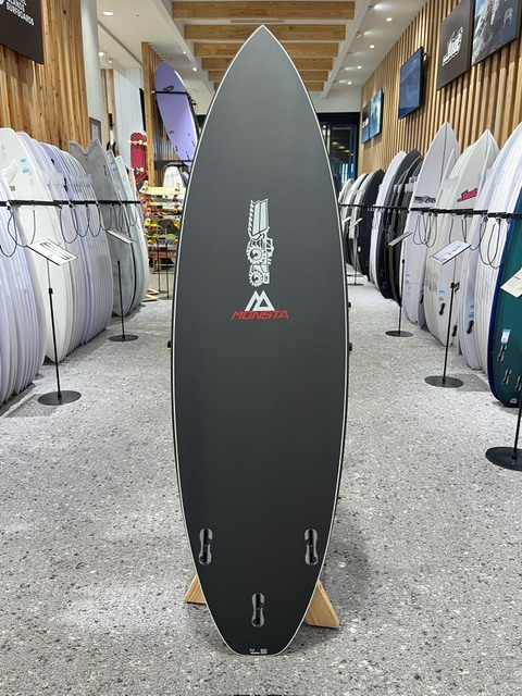 Tall black surfboard on display in a surf shop, Munsta logo in red visible on its surface.