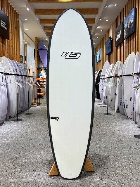 a-0 - SHOP NEWS | ムラサキスポーツオンラインストア 通販 Tall white surfboard with an HS logo stands upright in a surf shop showroom, surrounded by other boards on racks.