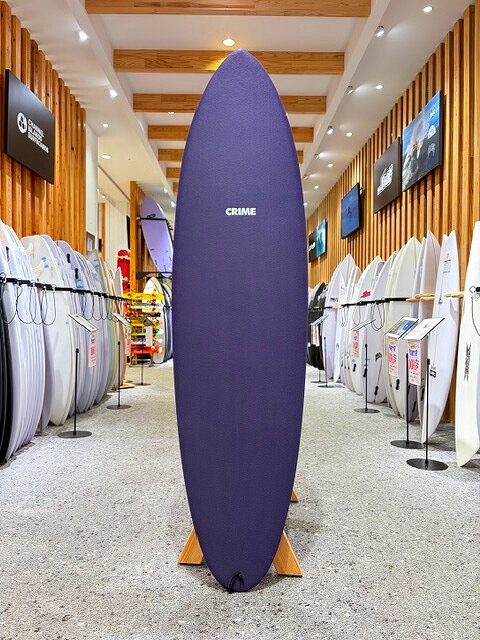 a-IMG_2140 - SHOP NEWS | ムラサキスポーツオンラインストア 通販 Tall purple surfboard with 'CRIME' logo on a stand, in a surf shop aisle surrounded by white boards and wooden wall panels.