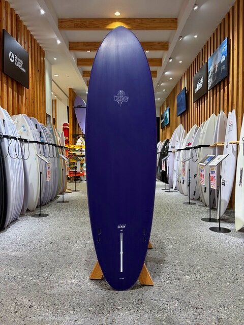 a-IMG_2141 - SHOP NEWS | ムラサキスポーツオンラインストア 通販 Tall blue surfboard on a wooden stand in a showroom, flanked by white surfboards along the walls.