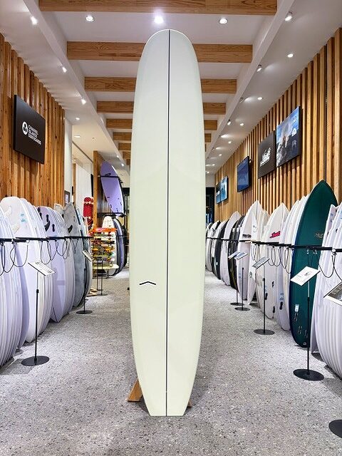a-IMG_2663 - SHOP NEWS | ムラサキスポーツオンラインストア 通販 Tall pale yellow surfboard standing upright on a stand in a surf shop, surrounded by rows of other boards on racks with wooden wall panels and ceiling lights.