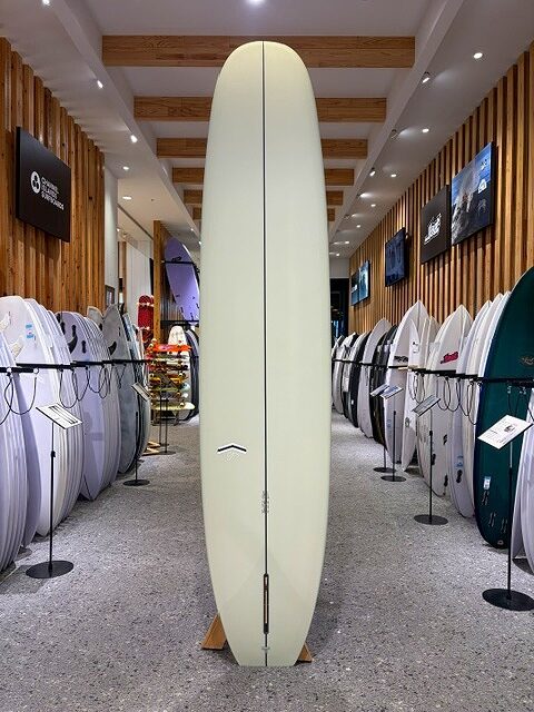 a-IMG_2664 - SHOP NEWS | ムラサキスポーツオンラインストア 通販 Tall pale-yellow surfboard standing upright on a wooden rack in a surf shop, flanked by rows of boards.