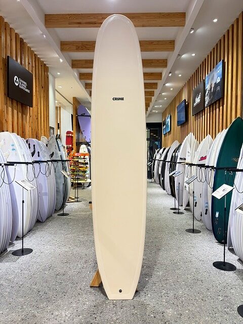 a-IMG_2665 - SHOP NEWS | ムラサキスポーツオンラインストア 通販 Cream surfboard on display in a showroom, centered among boards on wood-paneled walls; the center board reads CRIME.