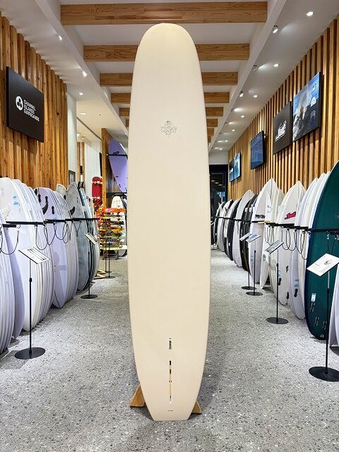 a-IMG_2666 - SHOP NEWS | ムラサキスポーツオンラインストア 通販 Tall cream-colored surfboard stands upright in the center of a surf shop, flanked by rows of boards on racks.