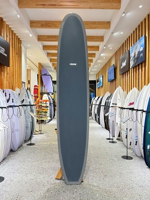 a-IMG_2667 - SHOP NEWS | ムラサキスポーツオンラインストア 通販 Tall gray surfboard standing upright on a display rack in a surf shop aisle.