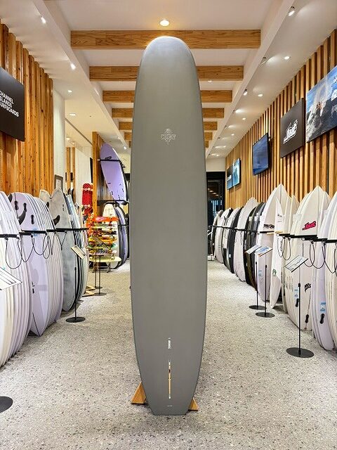 a-IMG_2668 - SHOP NEWS | ムラサキスポーツオンラインストア 通販 Indoor surf shop with a tall gray surfboard on a stand in the center, flanked by rows of white and colored boards along wooden wall displays.