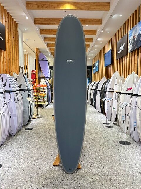 a-IMG_2670 - SHOP NEWS | ムラサキスポーツオンラインストア 通販 Tall dark gray surfboard on a stand in a showroom, surrounded by racks of surfboards along wood-paneled walls.