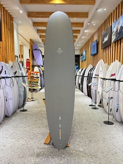 a-IMG_2671 - SHOP NEWS | ムラサキスポーツオンラインストア 通販 Gray surfboard standing upright on a wooden stand in a surf shop showroom, with many boards on display along the walls.