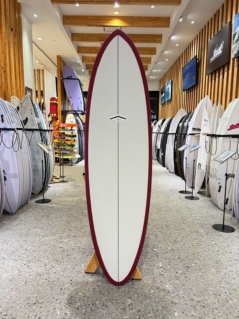 White surfboard with a burgundy rim on a display rack in a surf shop, surrounded by other boards on racks in a well-lit showroom.