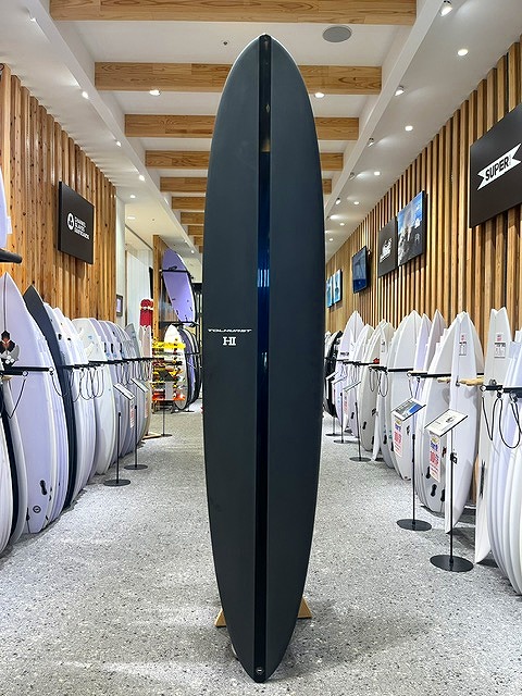 a-IMG_8596 - SHOP NEWS | ムラサキスポーツオンラインストア 通販 Tall dark blue surfboard standing upright in a surf shop showroom and flanked by other boards.
