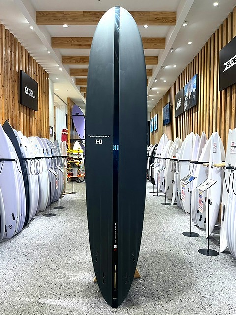 a-IMG_8598 - SHOP NEWS | ムラサキスポーツオンラインストア 通販 Tall dark blue surfboard standing upright in a surf shop aisle, with white boards on racks along both walls and wooden paneling visible.