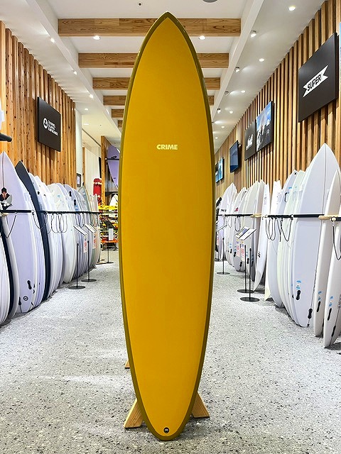 a-IMG_8666 - SHOP NEWS | ムラサキスポーツオンラインストア 通販 Yellow surfboard standing upright on a rack in a surf shop, surrounded by other boards on both sides.