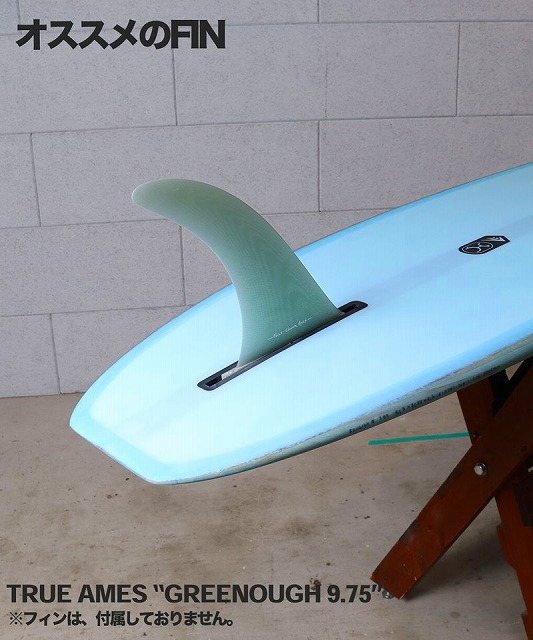 a-XTSbcLfg - SHOP NEWS | ムラサキスポーツオンラインストア 通販 Light blue surfboard with a pale green translucent fin attached, resting on a wooden stand against a gray wall.