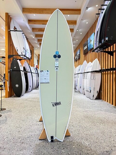 a-image0 - SHOP NEWS | ムラサキスポーツオンラインストア 通販 Cream-colored surfboard on a display stand inside a surf shop, with other boards visible on racks around it