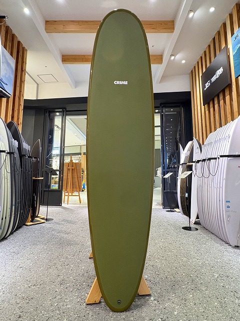 a-image0 - SHOP NEWS | ムラサキスポーツオンラインストア 通販 Olive green surfboard standing upright on a wooden rack inside a surf shop, with other boards displayed along the walls.