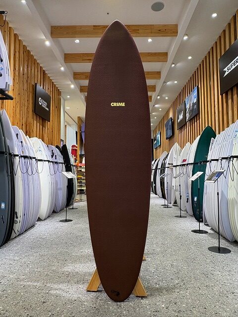 a-image0 - SHOP NEWS | ムラサキスポーツオンラインストア 通販 Brown surfboard displayed upright on a wooden stand in a surf shop, surrounded by racks of white and colored boards.