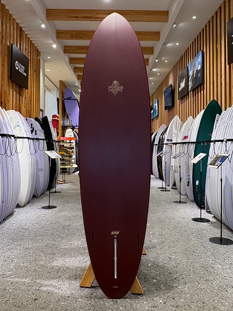 a-image0 - SHOP NEWS | ムラサキスポーツオンラインストア 通販 Tall maroon surfboard on a wooden stand in a bright surf shop, with white and colored boards lining both walls