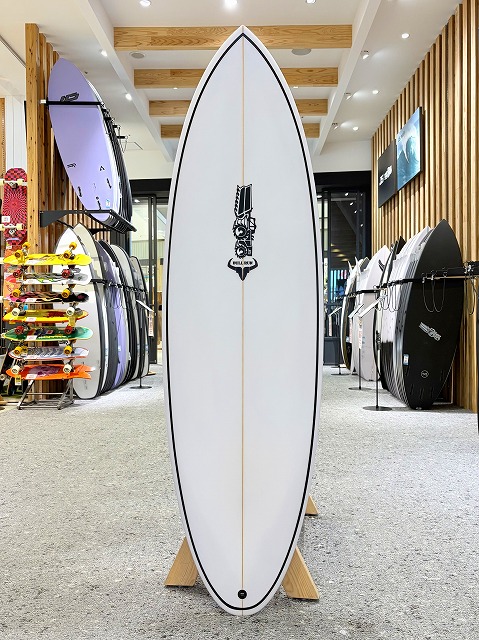 White surfboard on a wooden stand in a surf shop, with racks of colorful boards and gear visible in the background.