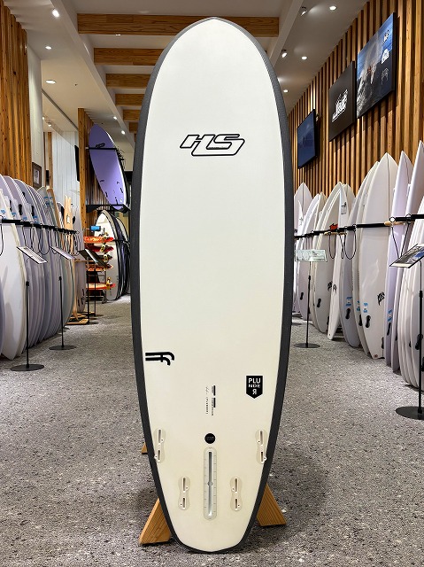 a-image1 - SHOP NEWS | ムラサキスポーツオンラインストア 通販 White surfboard with a black edge on a display rack in a surf shop, surrounded by many other boards on both sides.