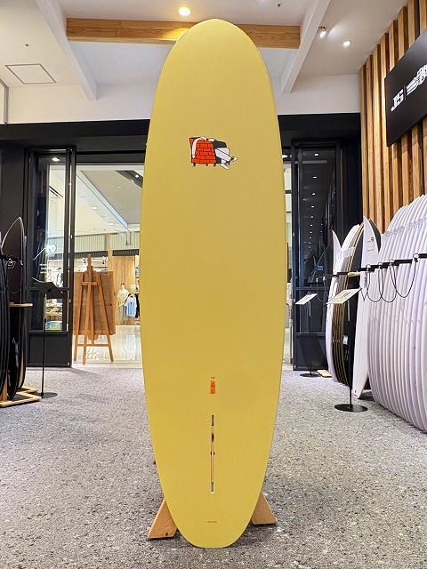 a-image1 - SHOP NEWS | ムラサキスポーツオンラインストア 通販 Yellow surfboard on a display stand in a surf shop, with other boards lined up to the right and store fixtures in the background.