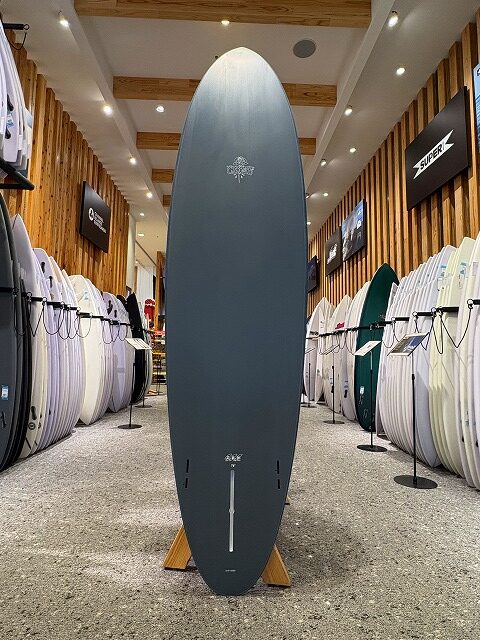 a-image1 - SHOP NEWS | ムラサキスポーツオンラインストア 通販 Tall dark gray surfboard standing upright on a wooden rack inside a surf shop, surrounded by numerous boards on both sides.