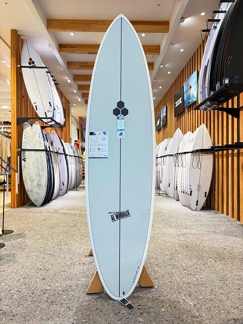 a-image10 - SHOP NEWS | ムラサキスポーツオンラインストア 通販 A white surfboard on a stand in a bright showroom, with rows of boards lining the walls behind it.