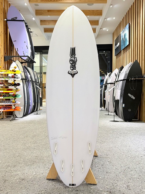 White surfboard on a wooden stand, upright in a surf shop showroom with other boards on racks along the walls.