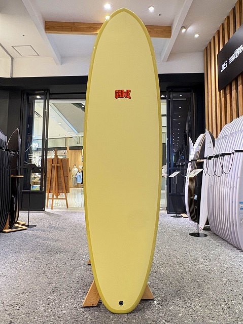 a-image2 - SHOP NEWS | ムラサキスポーツオンラインストア 通販 Yellow surfboard on a wooden stand in a showroom, with other boards and store interior visible, and a red 'CRIME' logo on the board.