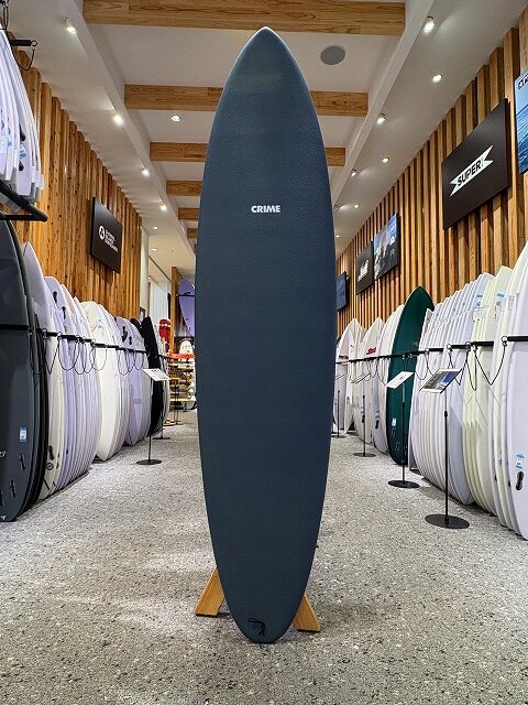 a-image2 - SHOP NEWS | ムラサキスポーツオンラインストア 通販 Tall dark blue surfboard on a wooden stand in a surf shop, with racks of white and colored boards on both sides and a visible 'CRIME' logo near the top.