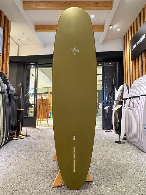 a-image3 - SHOP NEWS | ムラサキスポーツオンラインストア 通販 Olive-green surfboard standing upright on a wooden rack in a surf shop, with other boards visible on both sides.