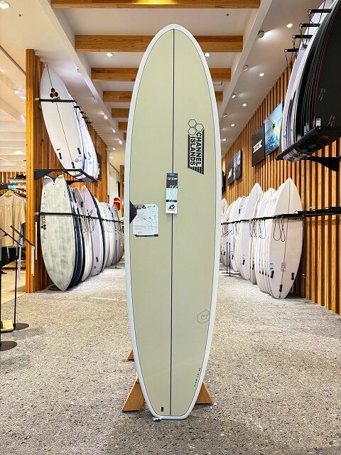 a-image4 - SHOP NEWS | ムラサキスポーツオンラインストア 通販 Beige longboard surfboard on a wooden display stand in a surf shop, with rows of boards lining the walls.