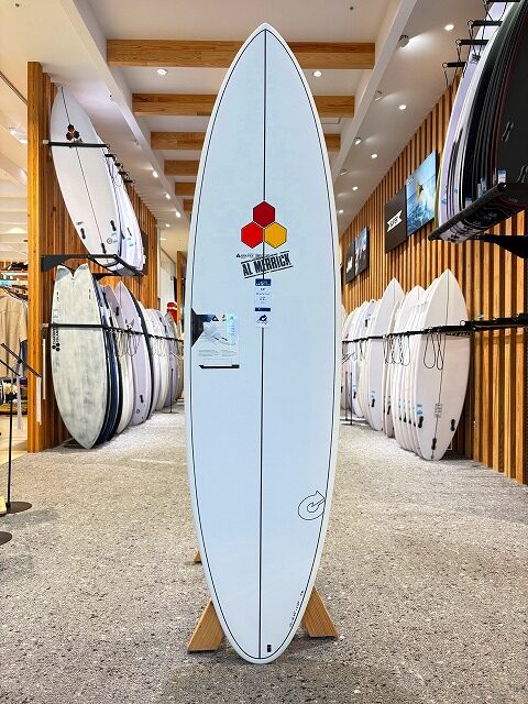 a-image5 - SHOP NEWS | ムラサキスポーツオンラインストア 通販 White surfboard on a wooden stand displayed upright in a surf shop, with racks of boards along the walls.