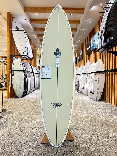 a-image7 - SHOP NEWS | ムラサキスポーツオンラインストア 通販 Cream-colored longboard on a display stand in a surf shop, with rows of surfboards lining both walls around it.