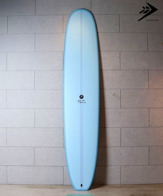 a-snmrBOJA - SHOP NEWS | ムラサキスポーツオンラインストア 通販 Light blue surfboard standing upright against a gray tiled wall; small black logo near center and a Firewire logo in the top-right corner.