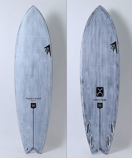 Two blue-gray surfboards with a weathered finish stand upright side by side; left shows the 'Seaside & Beyond' logo, right shows the 'Volcanic' logo and fin boxes at the tail.