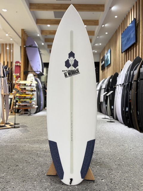 b-image0 - SHOP NEWS | ムラサキスポーツオンラインストア 通販 White surfboard on a stand inside a surf shop, with other boards and wooden wall accents in the background.