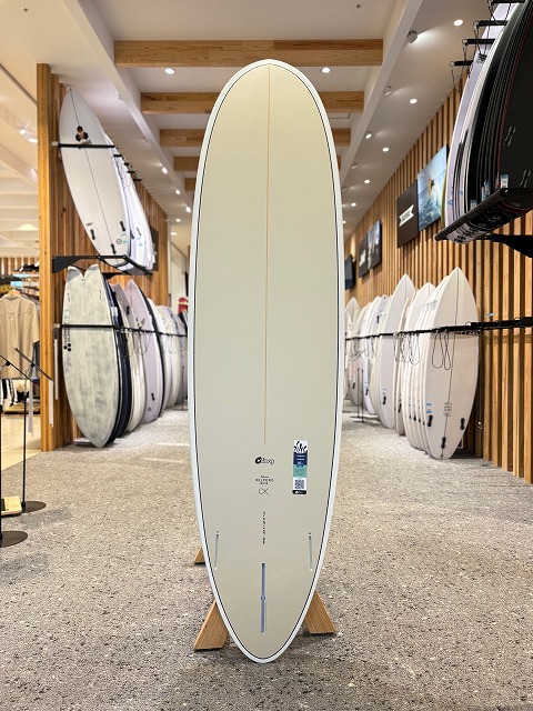 b-image0 - SHOP NEWS | ムラサキスポーツオンラインストア 通販 Tall, pale-yellow surfboard on a stand inside a surf shop, surrounded by racks of boards and wooden slat walls.