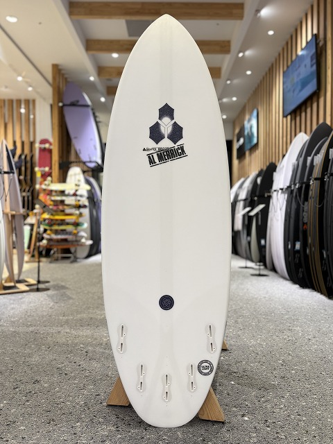 b-image1 - SHOP NEWS | ムラサキスポーツオンラインストア 通販 White surfboard on display in a surf shop, logo near the upper third and attached fins at the bottom, surrounded by other boards on racks.