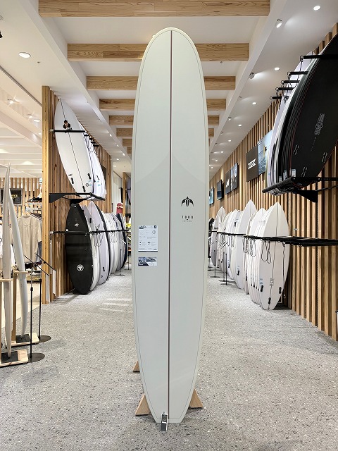 b-image2 - SHOP NEWS | ムラサキスポーツオンラインストア 通販 Tall white surfboard on a stand in a surf shop showroom with racks of boards on both sides.