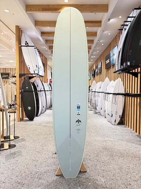 b-image5 - SHOP NEWS | ムラサキスポーツオンラインストア 通販 Pale mint-green surfboard on a display stand inside a surf shop, with racks of boards along both walls and wood paneling.