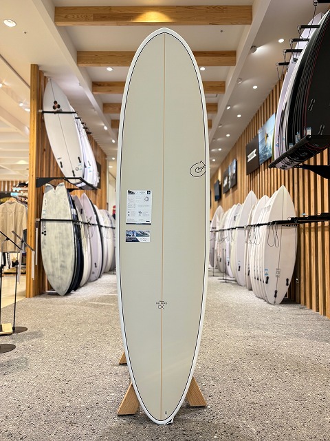 b-image6 - SHOP NEWS | ムラサキスポーツオンラインストア 通販 Cream-colored surfboard with a light stringer, standing upright on a display rack inside a bright surf shop.