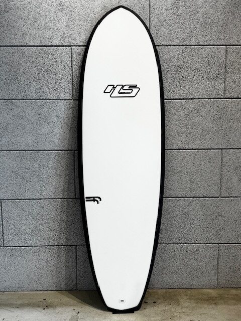 White surfboard with black rails standing upright against a gray brick wall; logos visible mid and lower sections.
