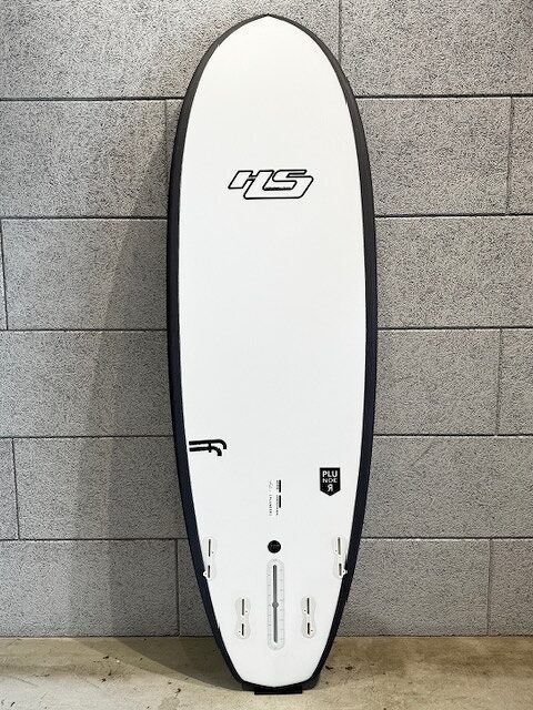 White surfboard with a black rim, standing upright against a gray tiled wall; features include an 'HS' logo near the top and multiple fin-box slots along the bottom edge.