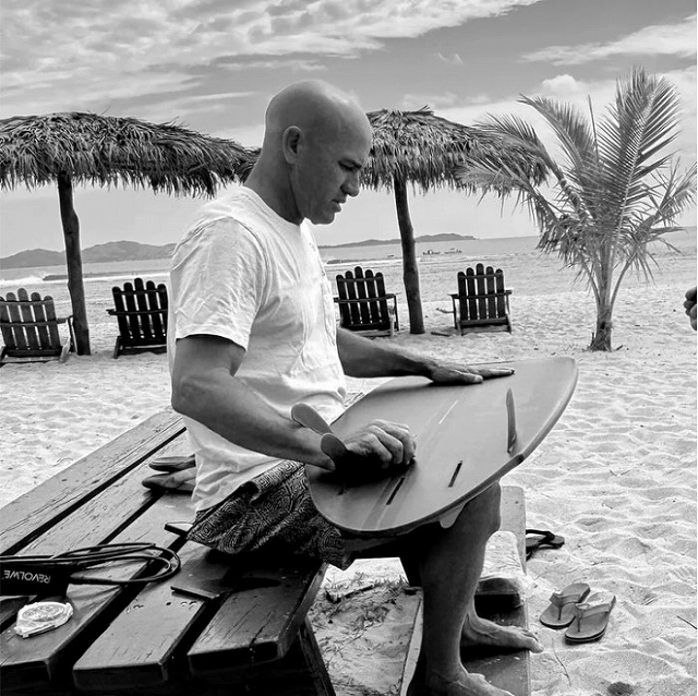 Bald man in a white t-shirt sits on a wooden bench on a beach, shaping a surfboard with a plane.