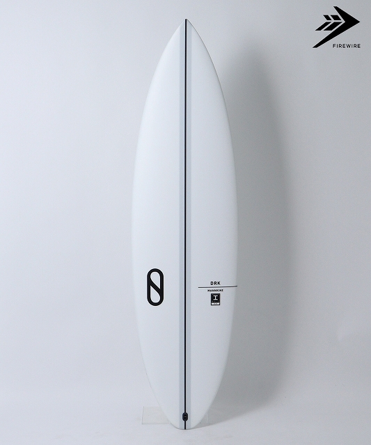 White surfboard standing upright against a pale wall, with a black logo on its left and a Firewire mark in the top-right corner.