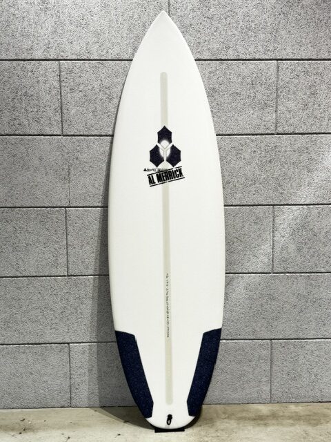 IMG_2860 - SHOP NEWS | ムラサキスポーツオンラインストア 通販 White surfboard standing upright against a gray brick wall; has a central fin box, dark blue tail pads, and a black hex logo near the top.