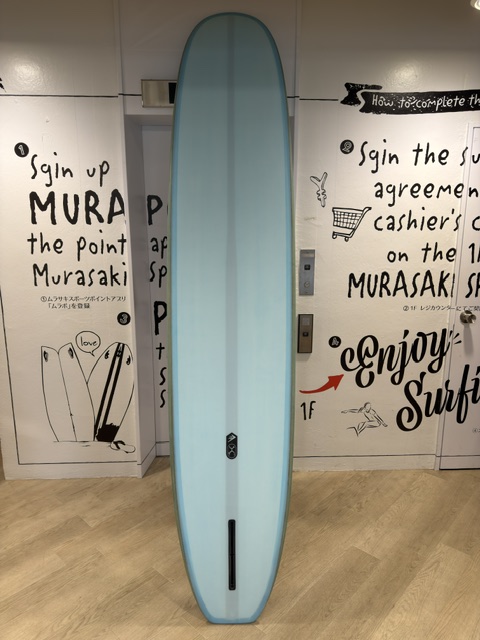 Tall light-blue surfboard standing upright indoors against white mural walls with drawings and text lines in the background, on a wooden floor.