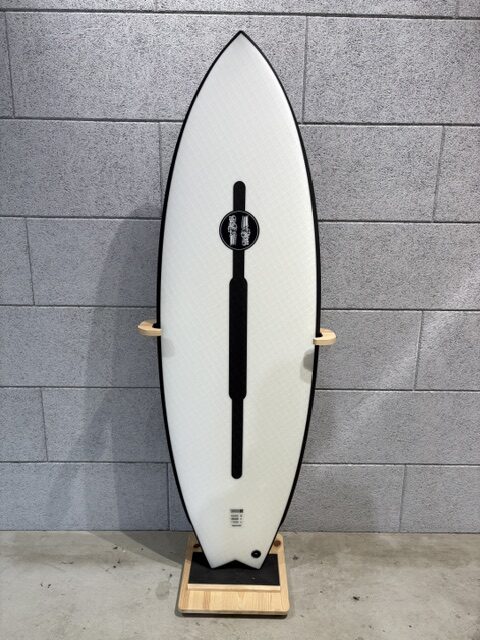 White surfboard with a black center channel standing upright on a wooden rack against a gray brick wall.