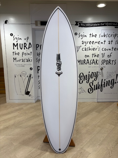 White surfboard with orange stringers on a display stand indoors, with posters in the background.
